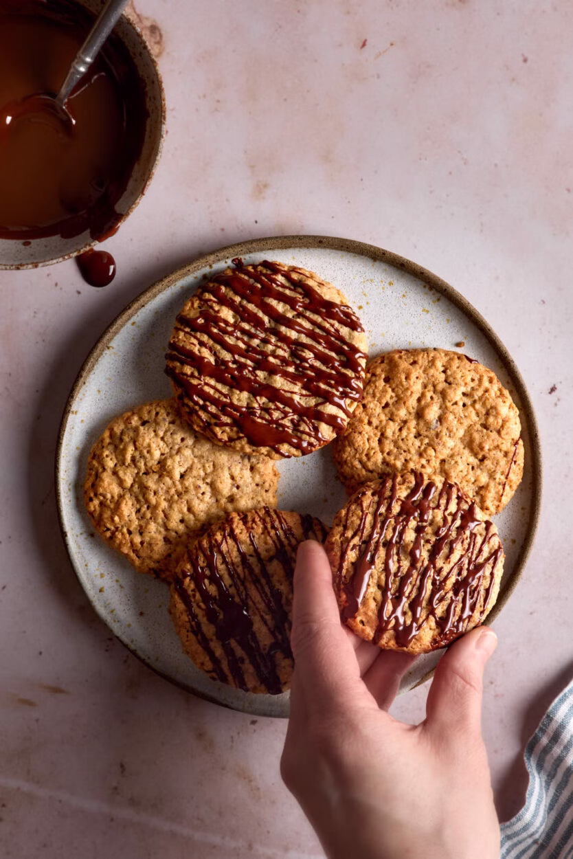 Ein Teller mit Hafercookies, teils mit Schokolade verziert. Eine Hand greift nach einem Keks, während eine Schüssel mit geschmolzener Schokolade danebensteht.