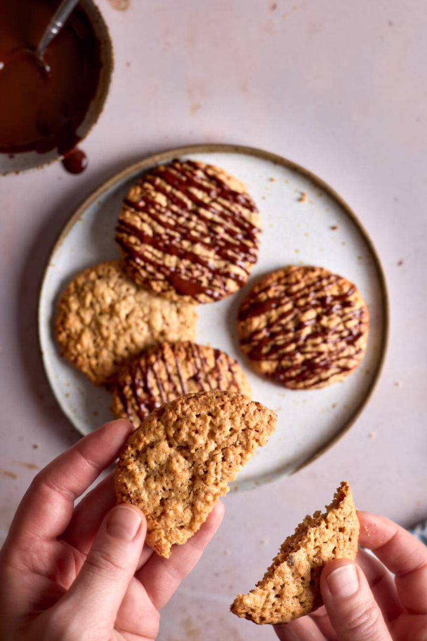 Zwei Hände brechen einen Haferkeks in der Mitte durch. Auf dem Teller dahinter liegen weitere Schwedische Hafercookies mit Schokodekor.