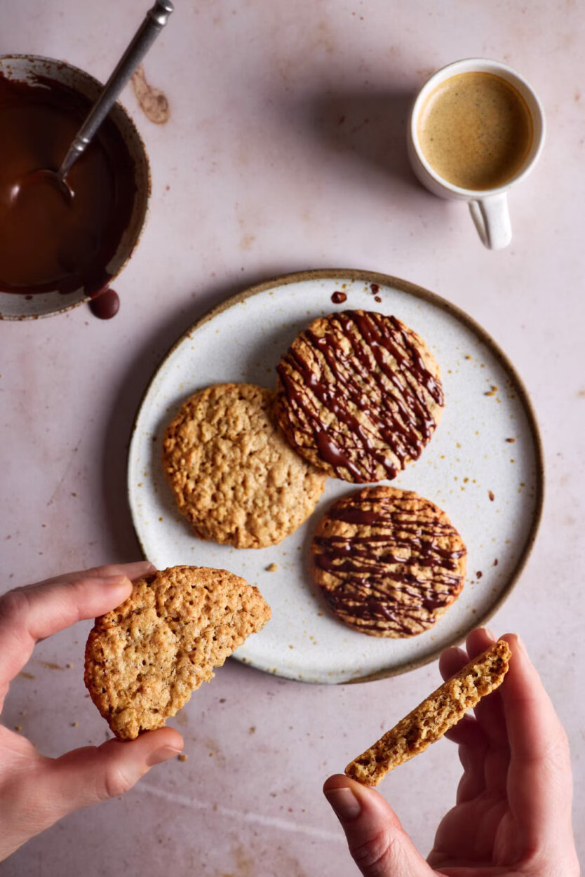 Zwei Hälften eines Haferkekses werden in die Kamera gehalten, während auf einem Teller weitere Hafercookies und eine kleine Espressotasse zu sehen sind.
