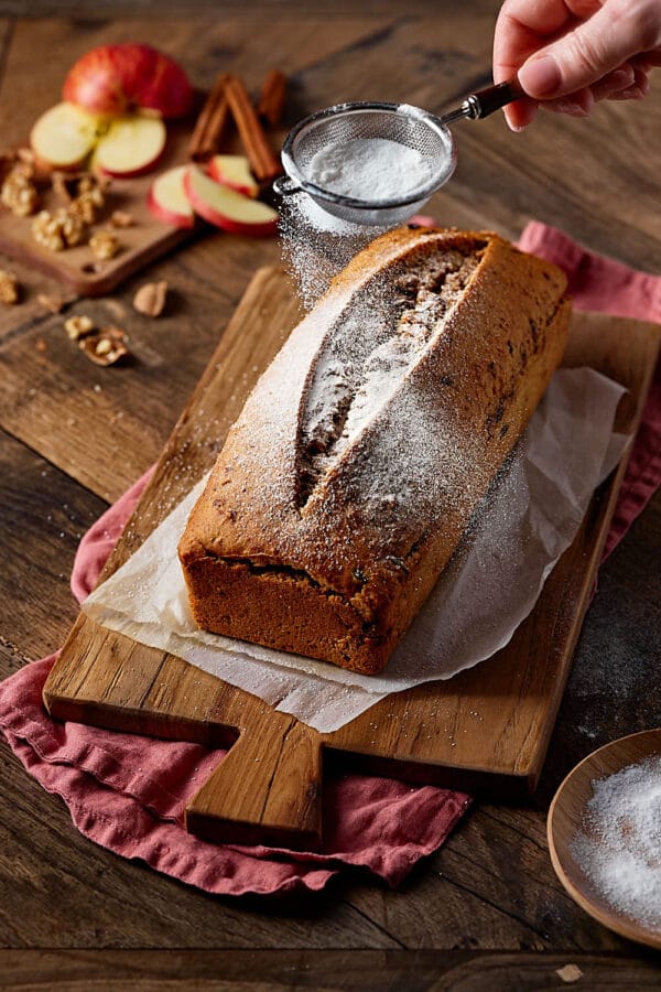 Saftiges Apfelbrot mit Nüssen, auf einem Holzbrett, das mit Puderzucker bestäubt wird.