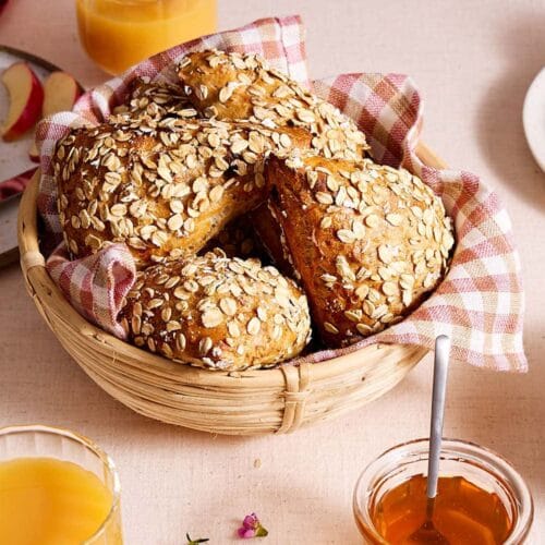 Saftige Müslibrötchen mit Quark in einem Brötchenkorb auf einem Frühstückstisch mit Orangensaft, Honig, Joghurt und aufgeschnittenem Apfel.