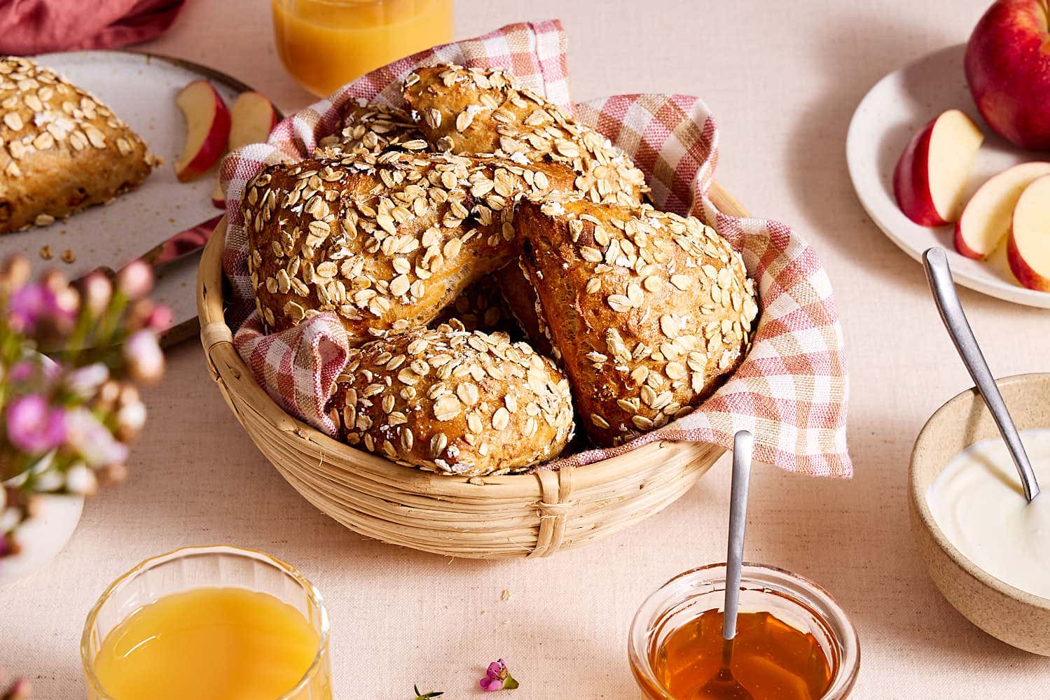 Saftige Müslibrötchen mit Quark in einem Brötchenkorb auf einem Frühstückstisch mit Orangensaft, Honig, Joghurt und aufgeschnittenem Apfel.