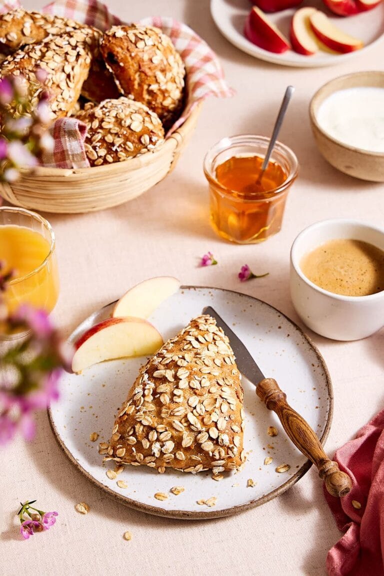 Müslibrötchen auf einem Teller, im Hintergrund Müslibrötchen in einem Brötchenkorb auf einem Frühstückstisch mit Orangensaft, Honig, Joghurt und aufgeschnittenem Apfel.