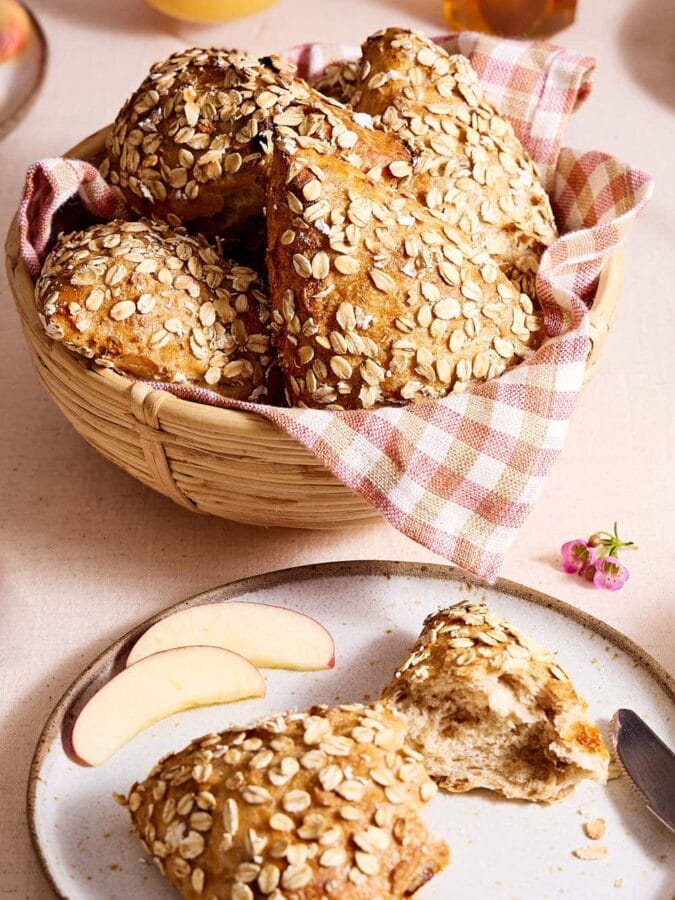 Müslibrötchen auf einem Teller, im Hintergrund Müslibrötchen in einem Brötchenkorb auf einem Frühstückstisch.