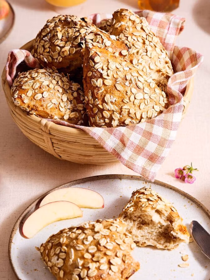 Müslibrötchen auf einem Teller, im Hintergrund Müslibrötchen in einem Brötchenkorb auf einem Frühstückstisch.