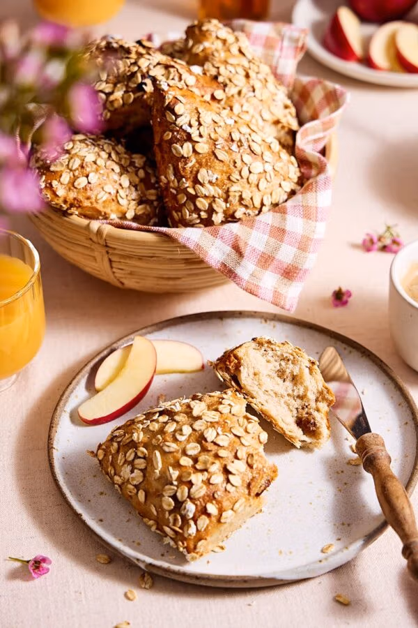 Müslibrötchen auf einem Teller, im Hintergrund Müslibrötchen in einem Brötchenkorb auf einem Frühstückstisch mit Orangensaft, Honig, Joghurt und aufgeschnittenem Apfel.