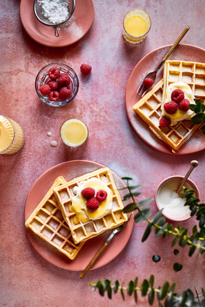 Eierlikörwaffeln mit Puderzucker, Sahne und frischen Himbeeren auf einem rosafarbenen Teller, umgeben von Puderzucker, einem Sieb und einem Glas Eierlikör.