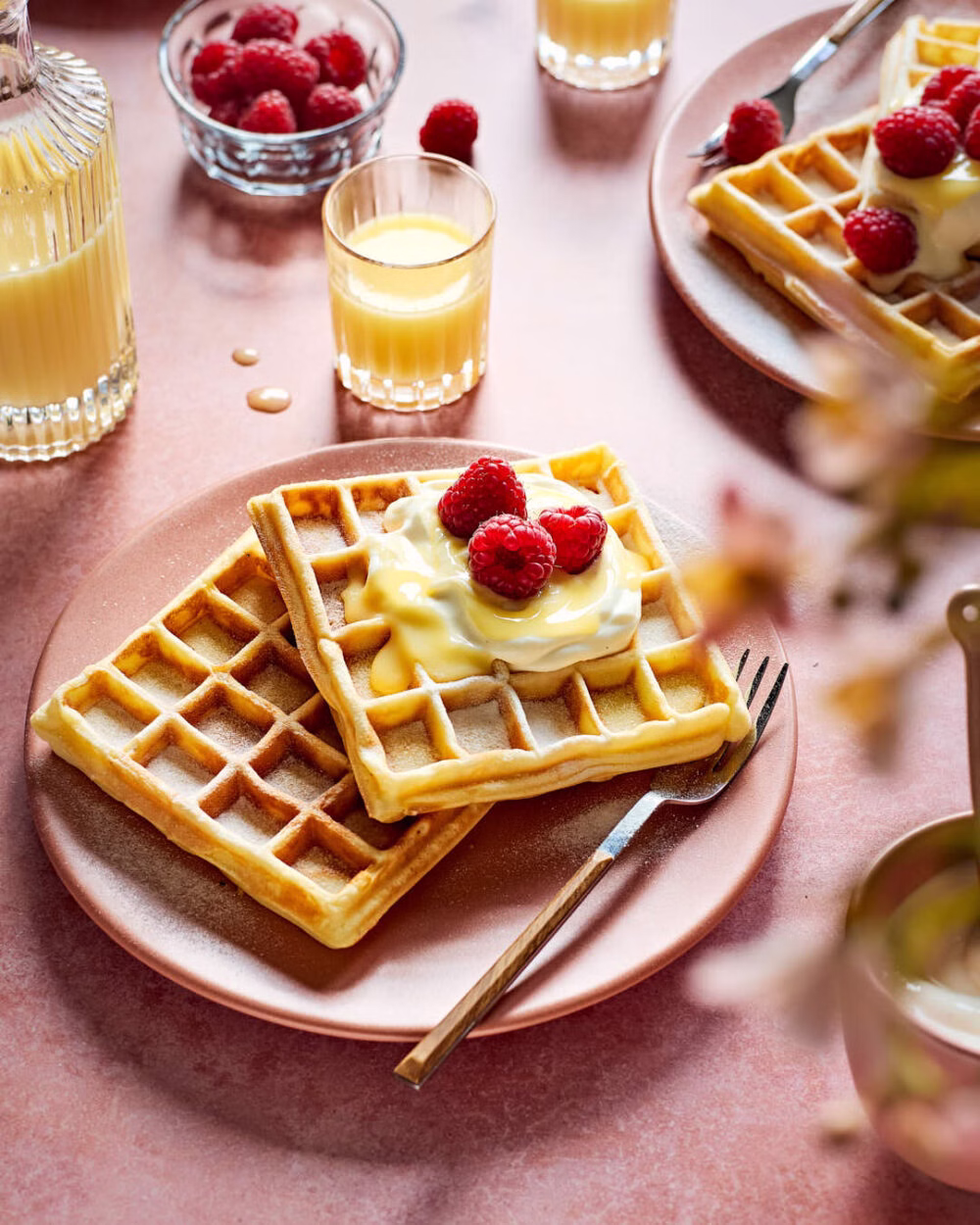 Goldbraune Belgische Waffeln mit Eierlikör, Sahne und frischen Himbeeren, serviert auf einem rosafarbenen Teller mit Gabel, umgeben von weiteren Waffeln, einem Glas Eierlikör und Beeren.