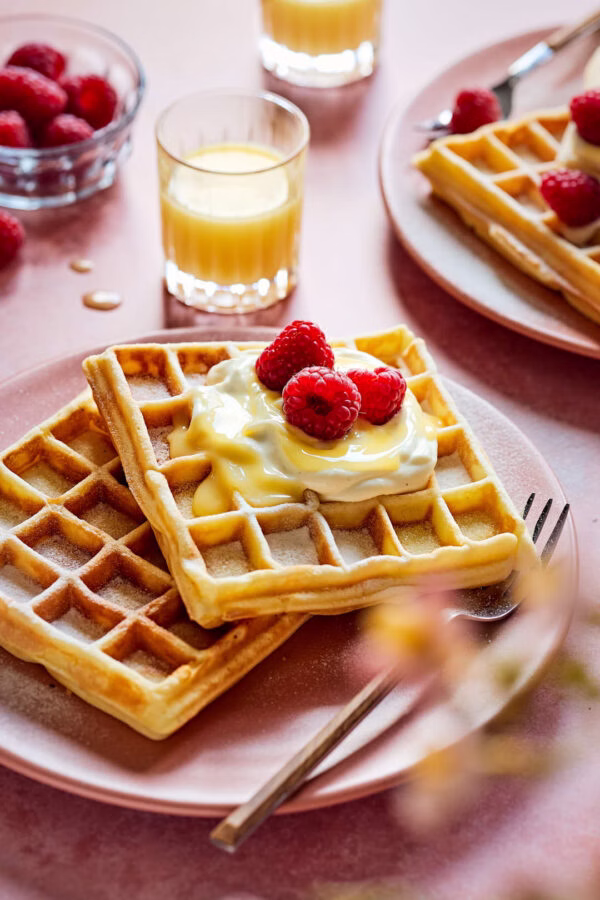 Goldbraune Belgische Waffeln mit Eierlikör, Sahne und frischen Himbeeren, serviert auf einem rosafarbenen Teller mit Gabel, umgeben von weiteren Waffeln, einem Glas Eierlikör und Beeren.