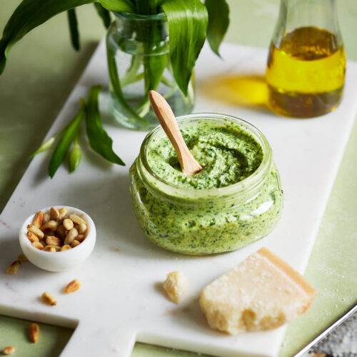 Ein Glas mit cremigem Bärlauch-Frischkäse-Pesto, umgeben von Pinienkernen, Parmesan und Olivenöl auf einem weißen Schneidebrett. Im Hintergrund steht ein kleines Glas mit frischem Bärlauch.