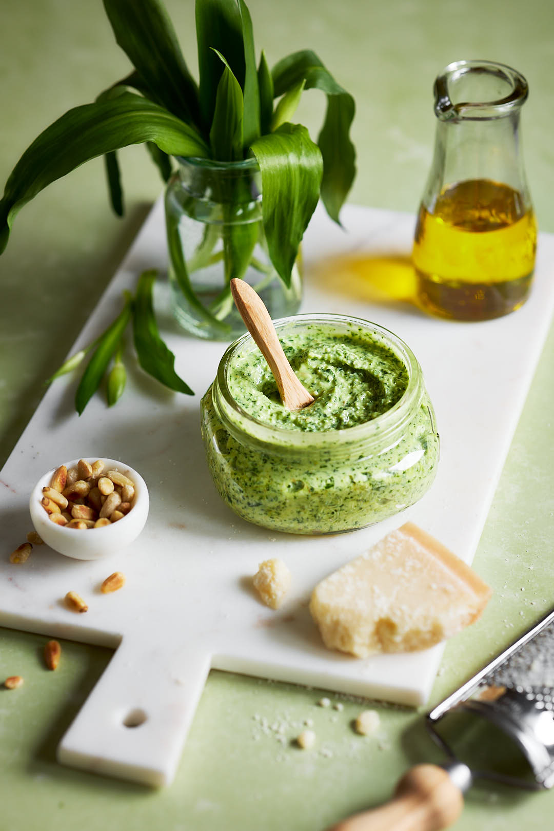 Ein Glas mit cremigem Bärlauch-Frischkäse-Pesto, umgeben von Pinienkernen, Parmesan und Olivenöl auf einem weißen Schneidebrett. Im Hintergrund steht ein kleines Glas mit frischem Bärlauch.