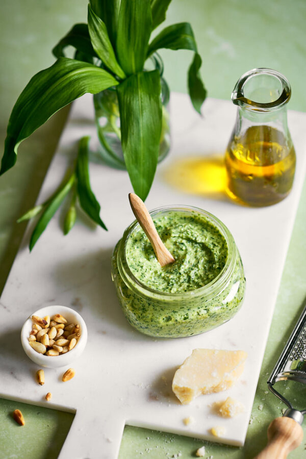Ein Glas mit grünem Bärlauch-Frischkäse-Pesto. Daneben liegen Pinienkerne, ein Stück Parmesan und ein Fläschchen Olivenöl. Frischer Bärlauch in einem Glas verleihen dem Bild eine frühlingshafte Note.