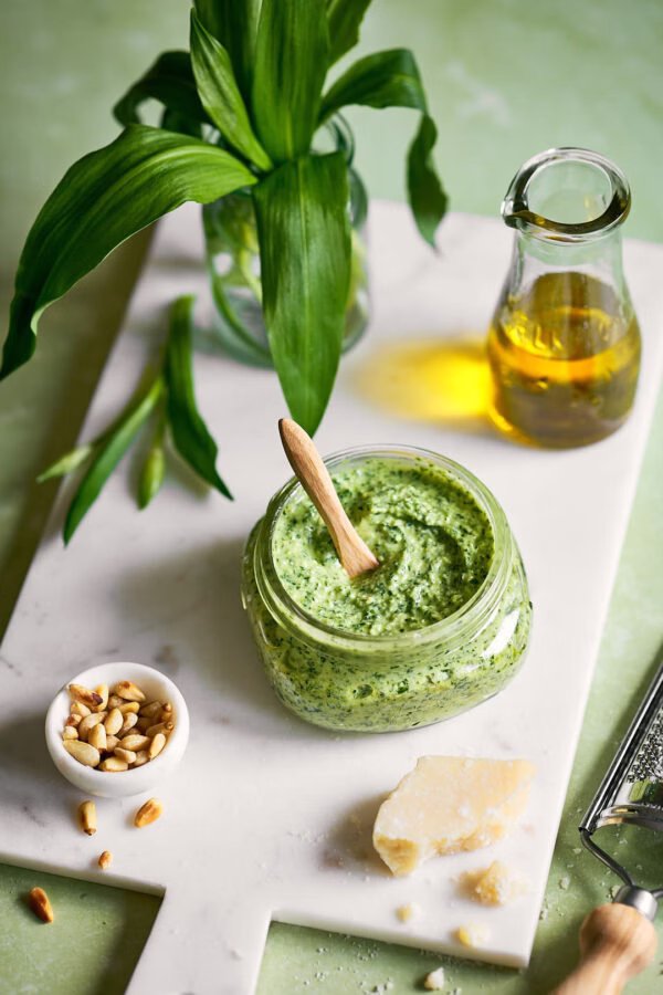 Ein Glas mit grünem Bärlauch-Frischkäse-Pesto. Daneben liegen Pinienkerne, ein Stück Parmesan und ein Fläschchen Olivenöl. Frischer Bärlauch in einem Glas verleihen dem Bild eine frühlingshafte Note.