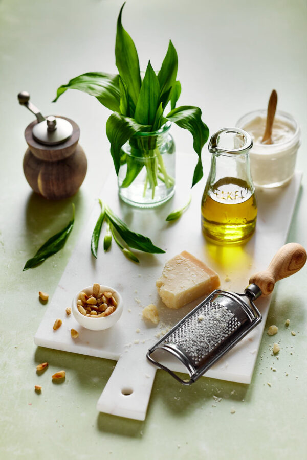 Zutaten für Bärlauch-Frischkäse-Pesto auf einem weißen Schneidebrett: frische Bärlauchblätter in einem Glas, Olivenöl, geriebener Parmesan, Pinienkerne und ein Pfeffermühle. Eine Reibe und ein Löffel Joghurt oder Frischkäse runden das Arrangement ab.