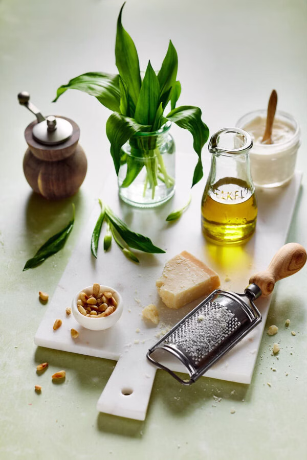 Zutaten für Bärlauch-Frischkäse-Pesto auf einem weißen Schneidebrett: frische Bärlauchblätter in einem Glas, Olivenöl, geriebener Parmesan, Pinienkerne und ein Pfeffermühle. Eine Reibe und ein Löffel Joghurt oder Frischkäse runden das Arrangement ab.