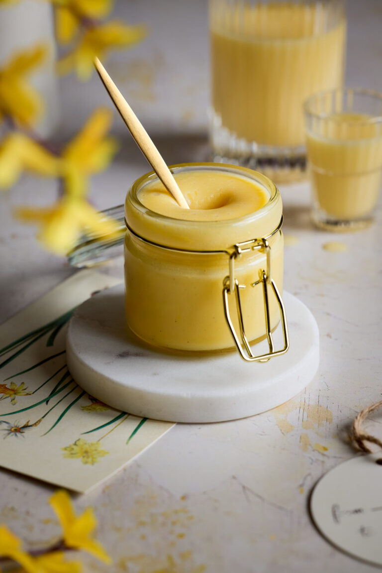 Cremige Eierlikörcreme mit einem goldenen Löffeln in einem Glas auf einem weißen Marmor-Untersetzer, im Hintergrund Eierlikör in einem Glas.