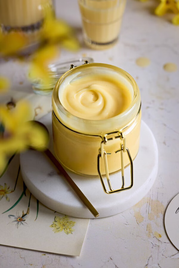Frische Eierlikörcreme im Glas, auf einer weißen Marmorplatte, umgeben von gelben Frühlingsblumen.