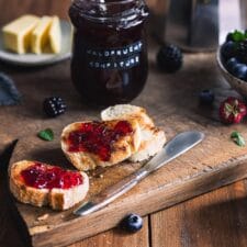 Waldfrucht Marmelade ohne Kerne auf gerösteten Baguettescheiben auf einem Holzbrett mit Beeren und Marmeladengläsern im Hintergrund.