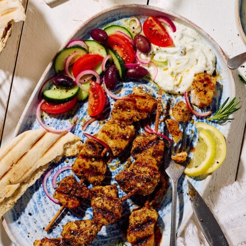 Marinierte Fleischspieße mit Zaziki, griechischem Bauernsalat, Zitronenscheiben und Pitabrot auf einer Servierplatte mit Weißwein im Hintergrund.