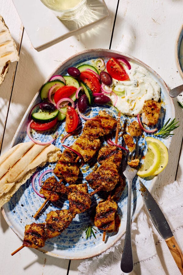 Marinierte Fleischspieße mit Zaziki, griechischem Bauernsalat, Zitronenscheiben und Pitabrot auf einer Servierplatte mit Weißwein im Hintergrund.
