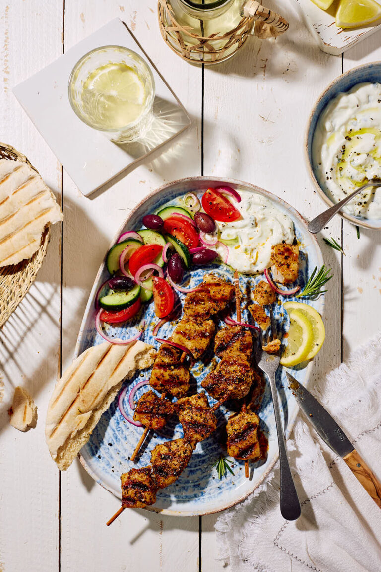 Marinierte Fleischspieße mit Zaziki, griechischem Bauernsalat, Zitronenscheiben und Pitabrot auf einer Servierplatte mit Weißwein im Hintergrund.