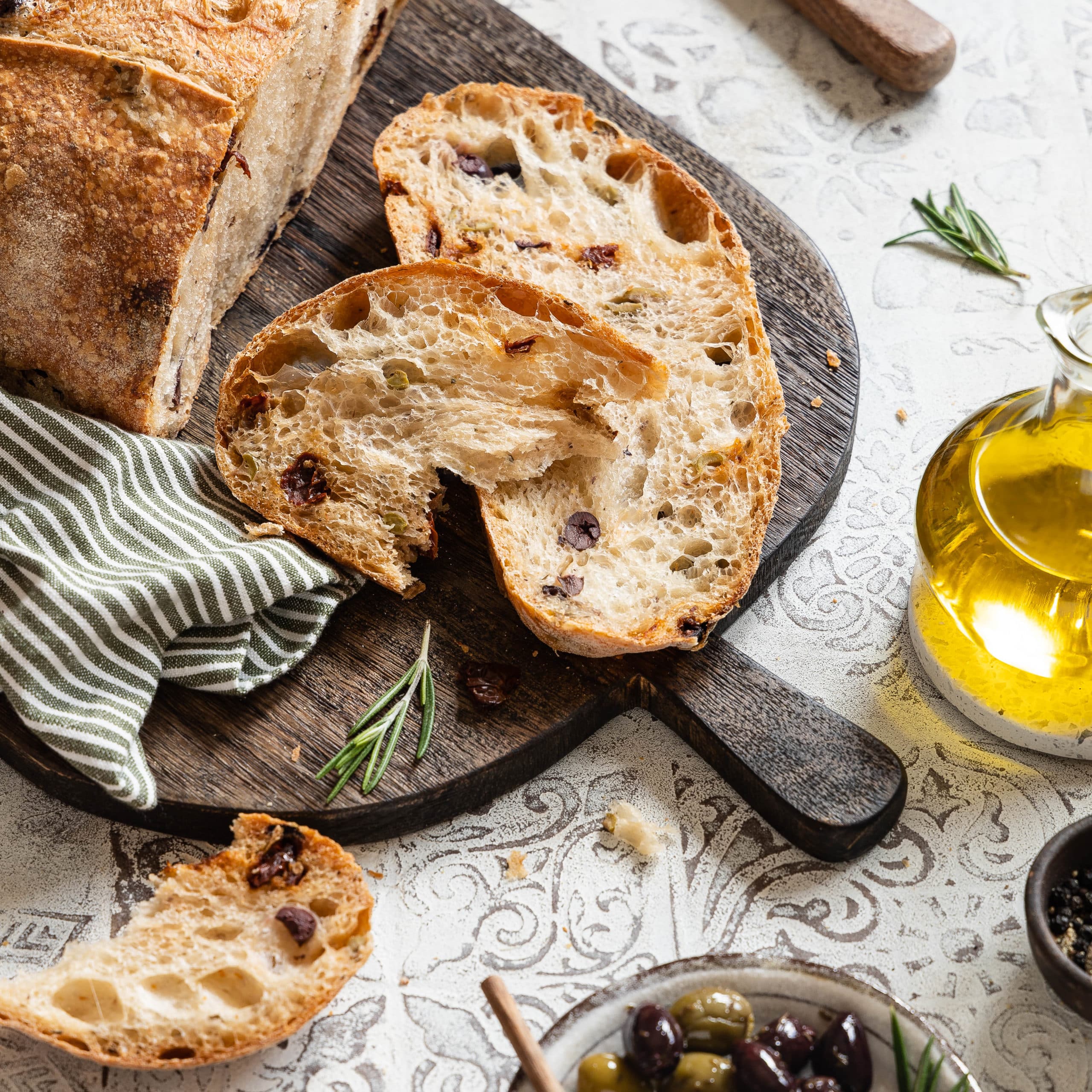 Mediterranes Olivenbrot in Scheiben geschnitten auf einem Holzbrett mit einer Schale Oliven und einer Glasflasche mit Olivenöl daneben.