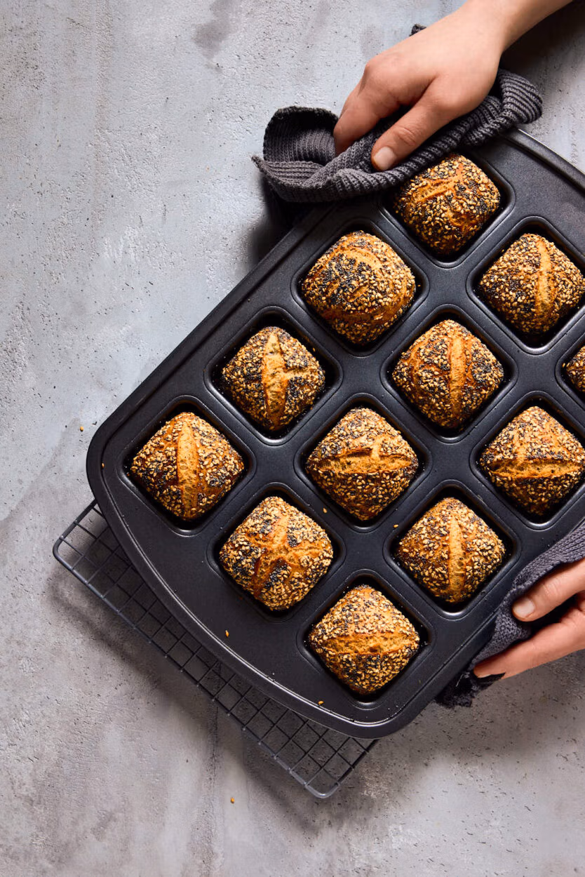 Zwei Hände heben eine Backform mit frisch gebackenen Mini-Weltmeisterbrötchen an. 