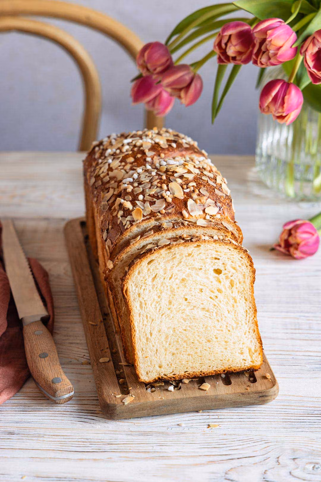 Nahaufnahme eines angeschnittenen Quarkstutens. Das fluffige Innere und die gerösteten Mandeln auf der Kruste sind deutlich zu erkennen. Im Hintergrund sind Tulpen zu sehen.