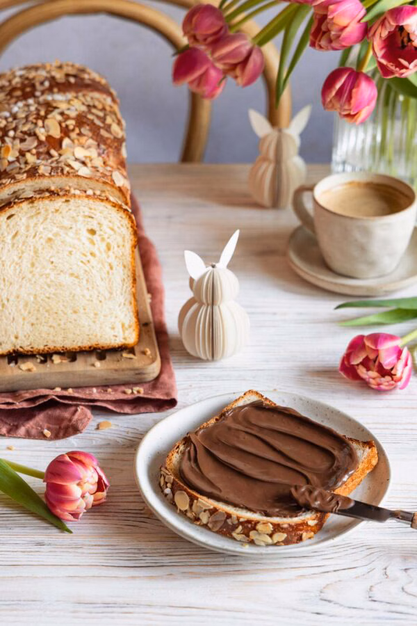 Eine Scheibe Quarkstuten mit Schokoladenaufstrich auf einem Teller. Daneben steht der angeschnittene Laib des lockeren Hefebrots mit Mandelsplittern. Im Hintergrund sind bunte Tulpen und eine Kaffeetasse zu sehen.