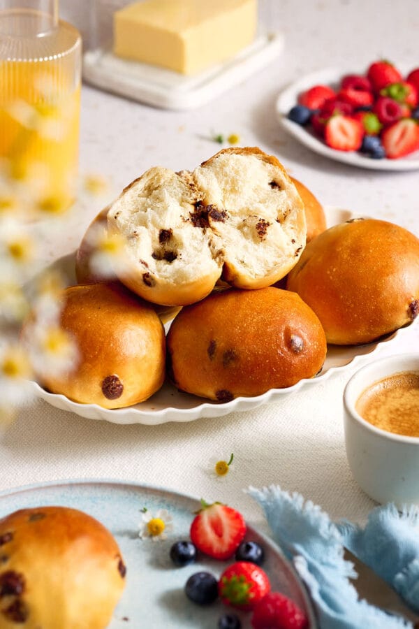 Schokobrötchen mit fluffiger Krume auf einem Teller, der in auf einem Frühstückstisch mit Obst, Orangensaft und Butter steht.