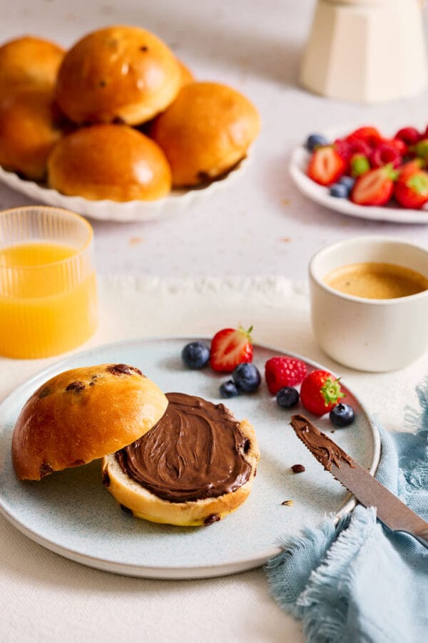 Fluffiges Schokobrötchen auf einem Teller mit Nuss-Nougat-Creme und einem gedeckten Frühstückstisch im Hintergrund. 