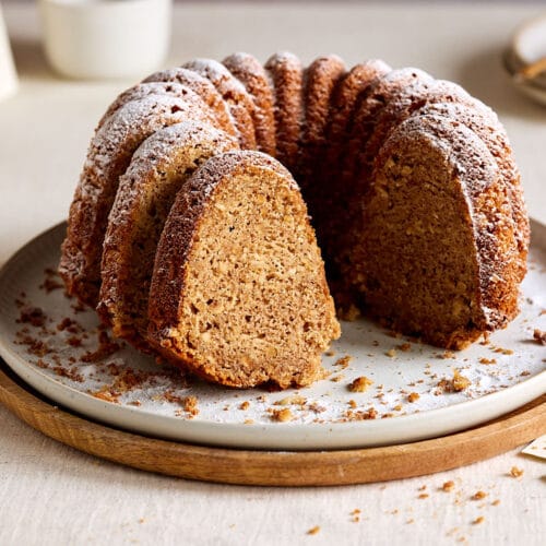 Saftiger Apfel-Dinkelkuchen, gebacken als Gugelhupf auf einem Teller mit Tassen und einer Kaffeekanne im Hintergrund.