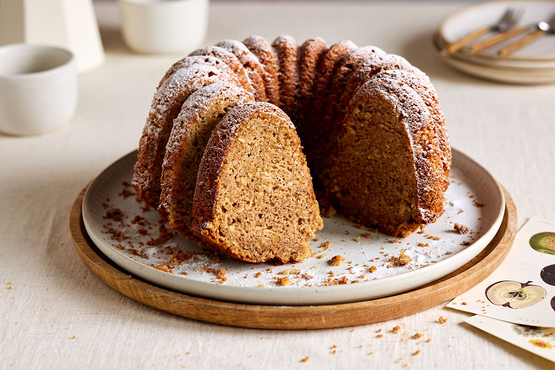 Saftiger Apfel-Dinkelkuchen, gebacken als Gugelhupf auf einem Teller mit Tassen und einer Kaffeekanne im Hintergrund.