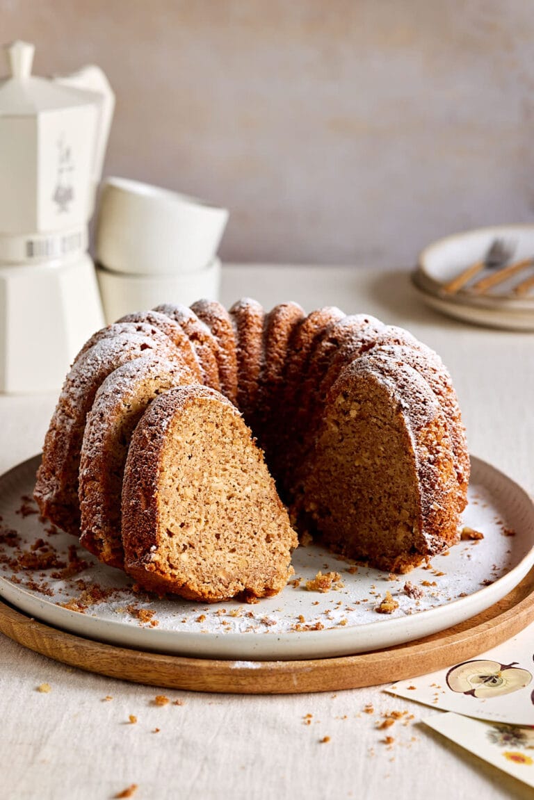 Saftiger Apfel-Dinkelkuchen, gebacken als Gugelhupf auf einem Teller mit Tassen und einer Kaffeekanne im Hintergrund.