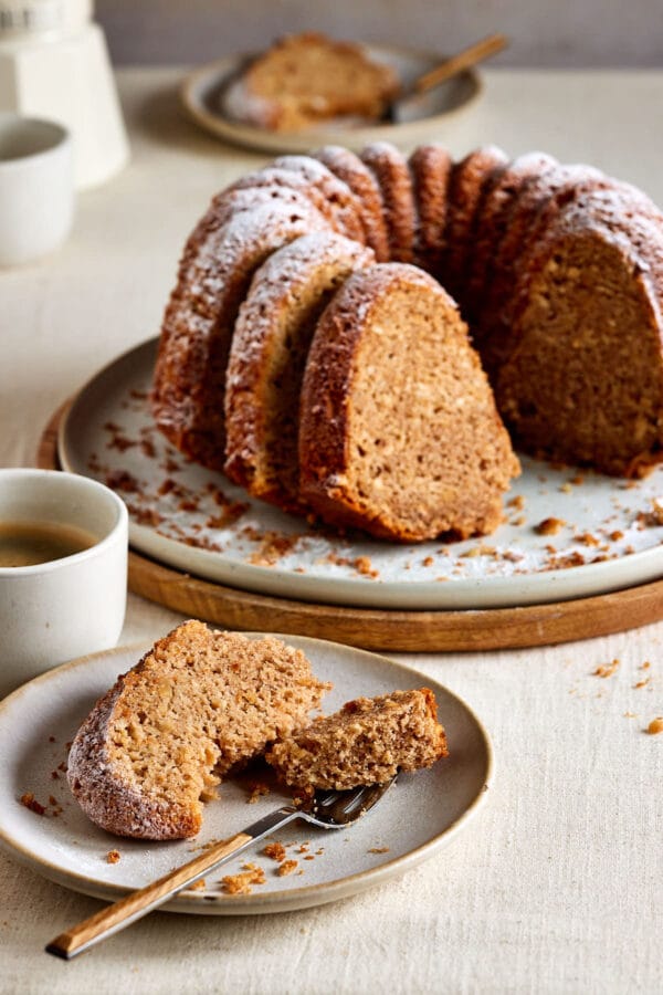 Saftiger Apfel-Dinkelkuchen, gebacken als Gugelhupf auf einem Teller mit Tassen und einer Kaffeekanne im Hintergrund. 