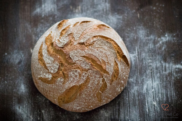 Ein runder Laib Dinkel-Bauernkruste mit knuspriger, bemehlter Oberfläche und einem dekorativen Muster auf dunklem Untergrund.