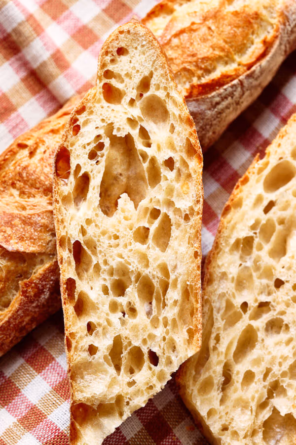 Nahaufnahme von einem französischen Baguette mit lockerer Krume, mit einem karierten Tuch auf einem rustikalen Holzbrett.