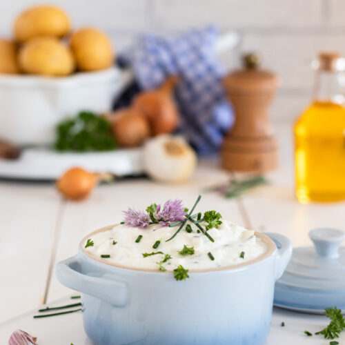 Kräuterquark mit Schnittlauch Petersilie und Schnittlauchblüten. Im Hintergrund Kartoffeln, Leinöl, Zwiebeln und Knoblauch sowie eine Pfeffermühle.