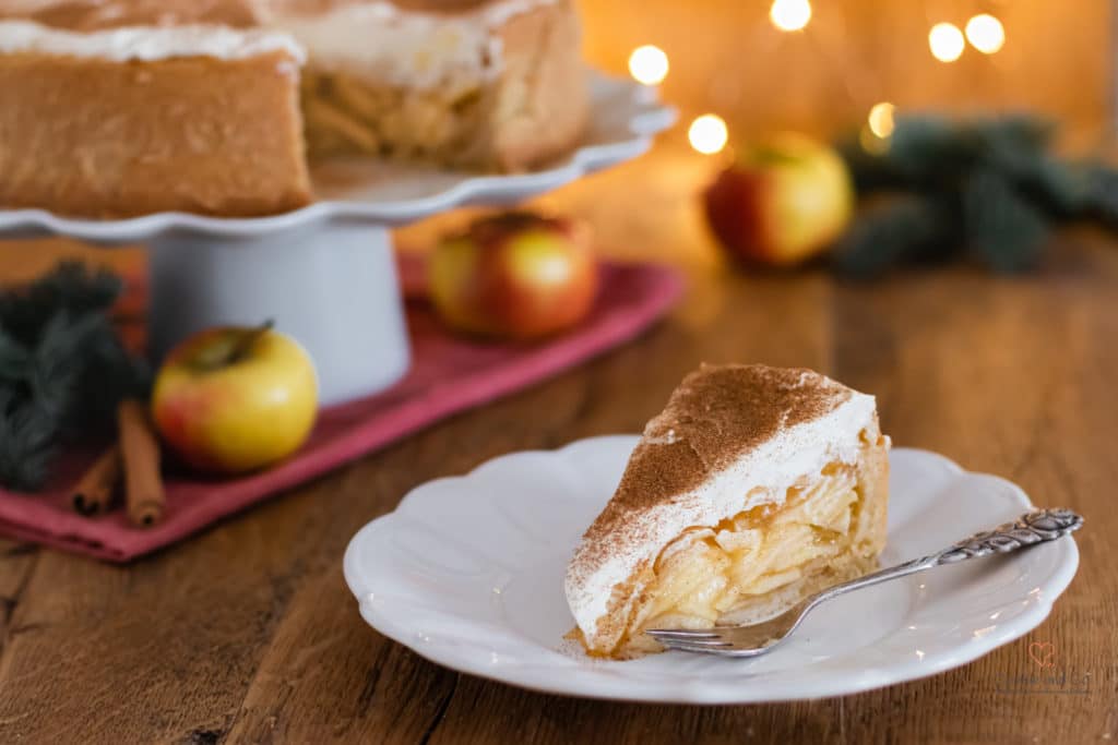 Kuchenstück von einer Apfel-Glögg-Torte (Apfel-Wein-Torte) auf einem Teller mit Weihnachtsbeleuchtung im Hintergrund.