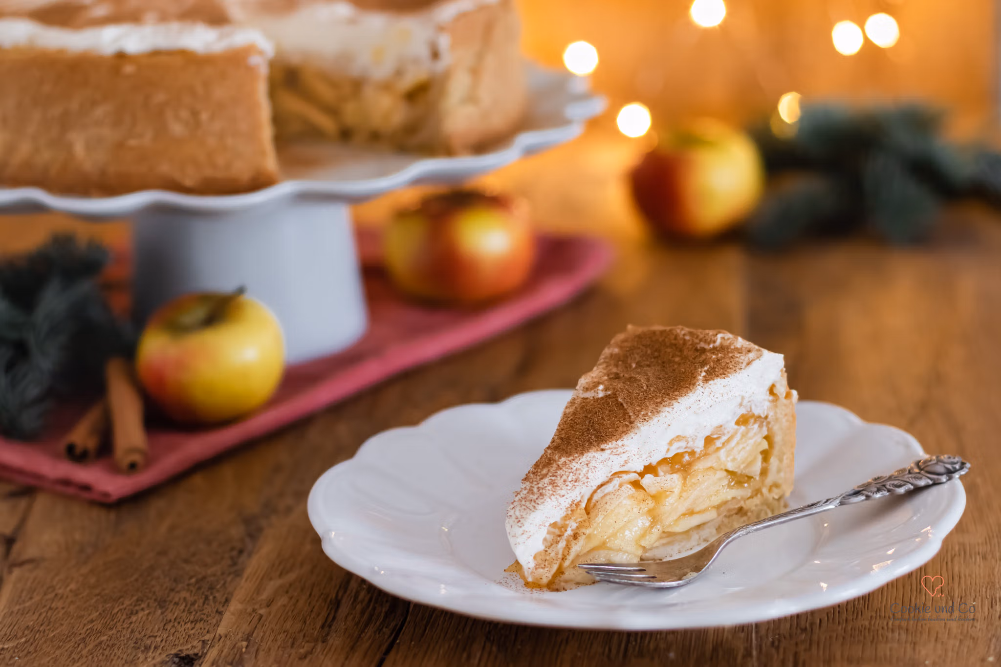 Ein Kuchenstück von einer Apfel-Glögg-Torte auf einem Teller mit weihnachtlicher Deko im Hintergrund.