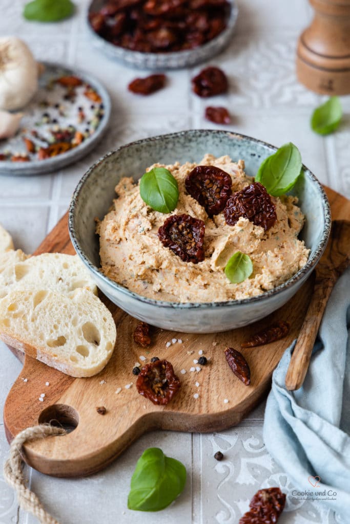 Mediterrane Tomaten-Feta-Creme in einer Schale auf einem Holzbrett mit Baguette und frischen Basilikumblättchen.