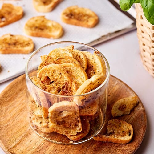 Selbstgemachte Brotchips in einem Glas mit einem Blech mit Brotchips im Hintergrund.