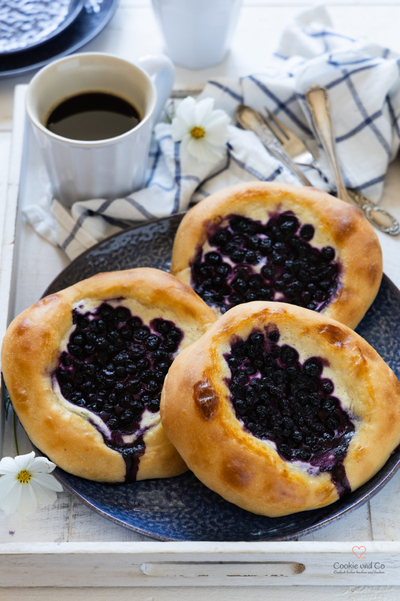 Blaubeer-Quark-Taler auf einem Teller mit einem weißen Tablett und einer Tasse Kaffee im Hintergrund.