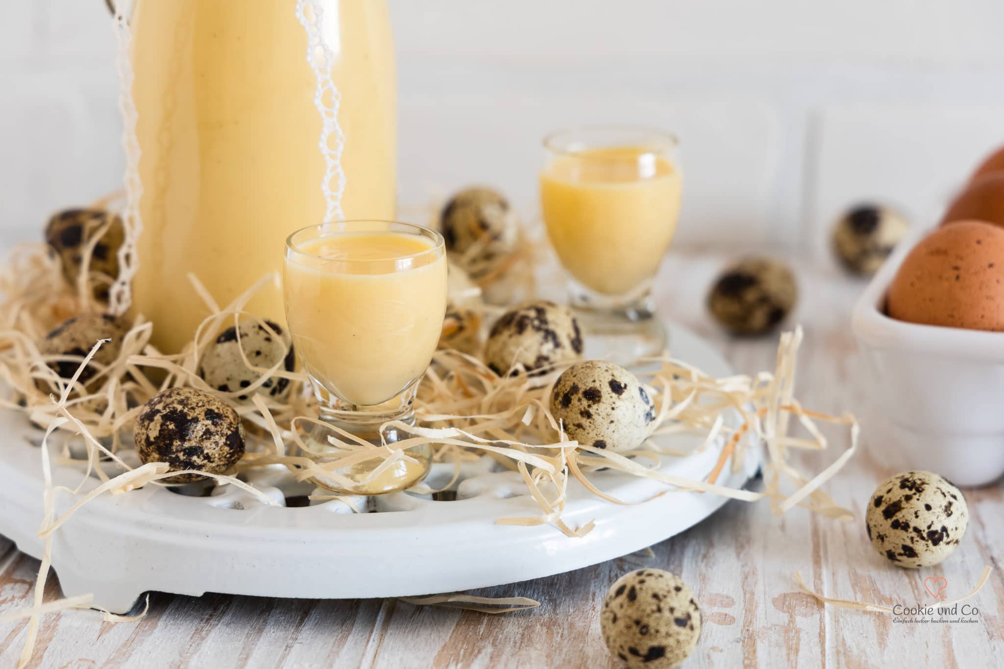 Selbstgemachter Eierlikör aus ganzen Eiern in einer dekorativen Glasflasche mit zwei gefüllten Likörgläsern, umgeben von Wachteleiern und Stroh auf einer weißen Servierplatte.