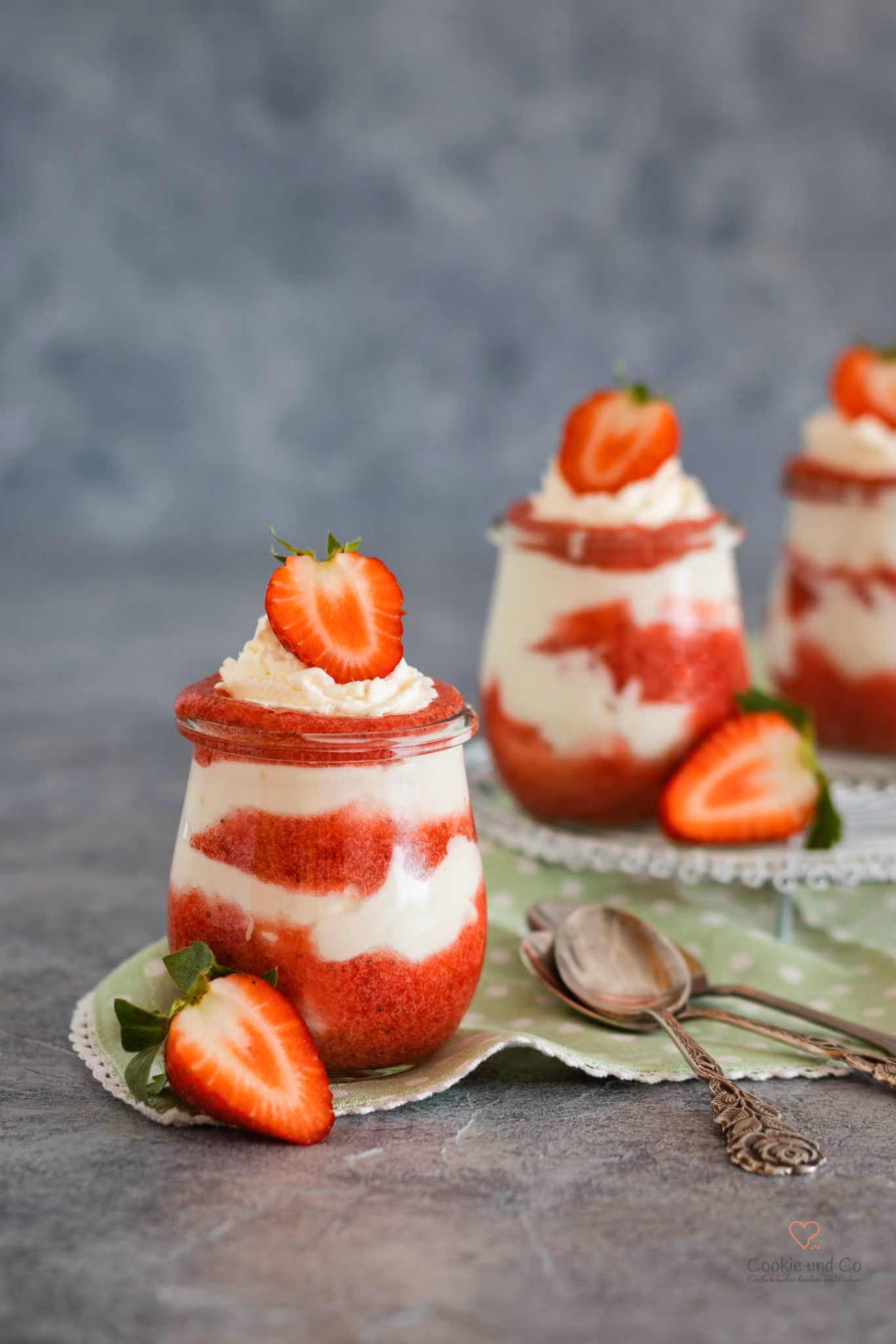 Erdbeer-Traum: In einem kleinen Glas geschichtetes Erdbeer-Sahne-Dessert mit frischen Erdbeerscheiben als Dekoration. Daneben liegen halbierte Erdbeeren und ein antiker Löffel auf einer grünen Serviette.