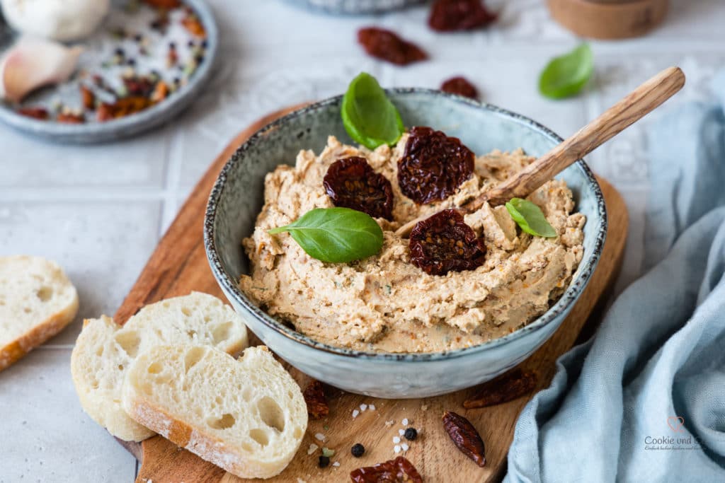 Mediterrane Tomaten-Feta-Creme in einer Schale auf einem Holzbrett mit Baguette und frischen Basilikumblättchen.