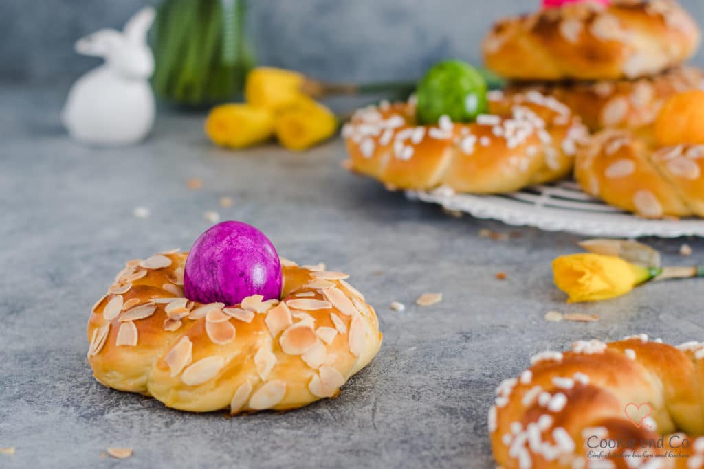 Osterkränze aus Dinkelmehl mit gefärbten Eiern und Narzissen im Hintergrund.