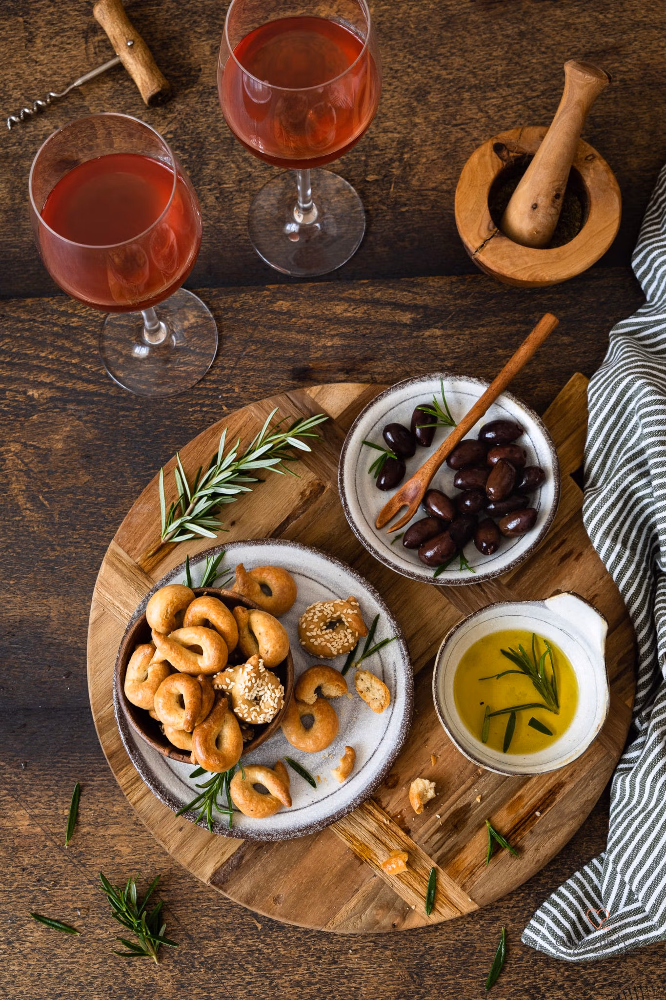 Taralli – ein italienisches Knabbergebäck auf einem Teller mit Olivenöl, Oliven, Rosmarin und Wein im Hintergrund.