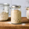Sauerteig auffrischen - Anstellgut in einem Glas auf einem rustikalen Holzbrett, mit Mehl und einem Wasserkrug im Hintergrund.