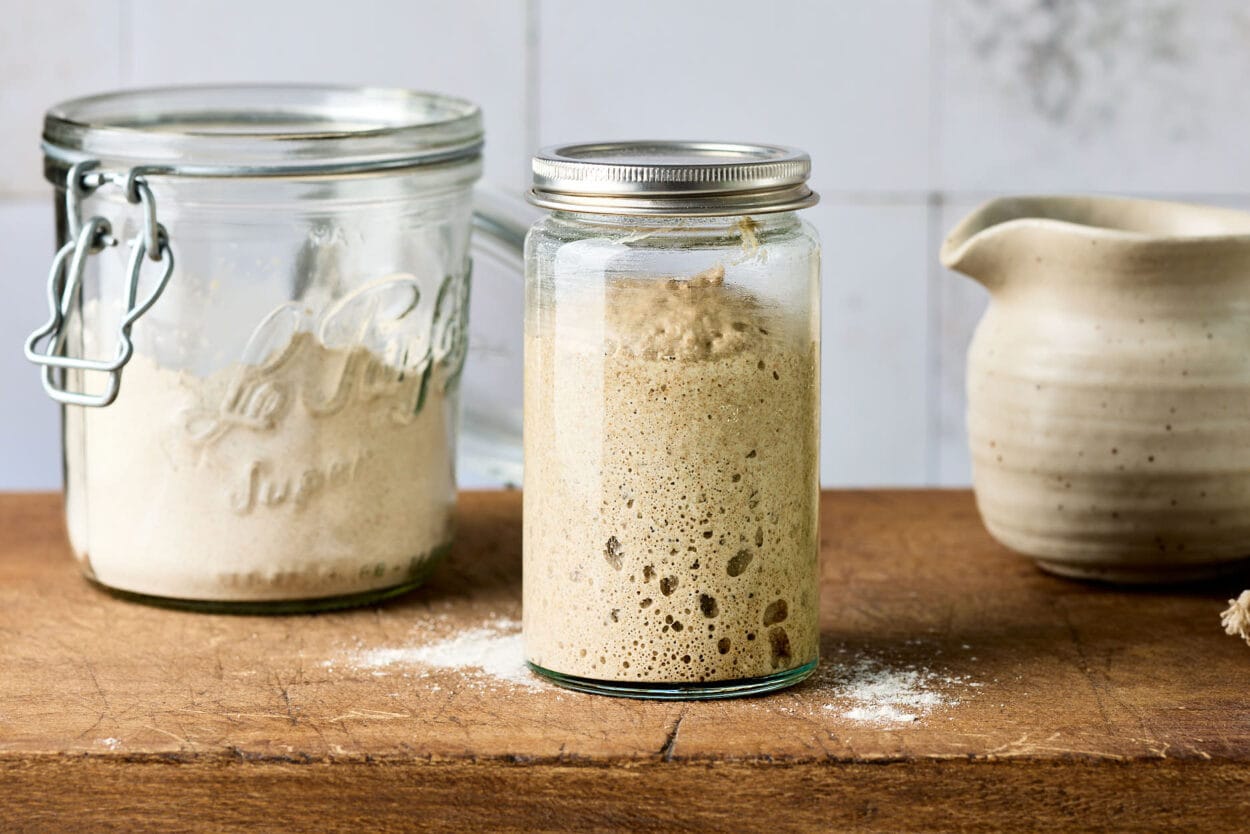 Sauerteig auffrischen - Anstellgut in einem Glas auf einem rustikalen Holzbrett, mit Mehl und einem Wasserkrug im Hintergrund.